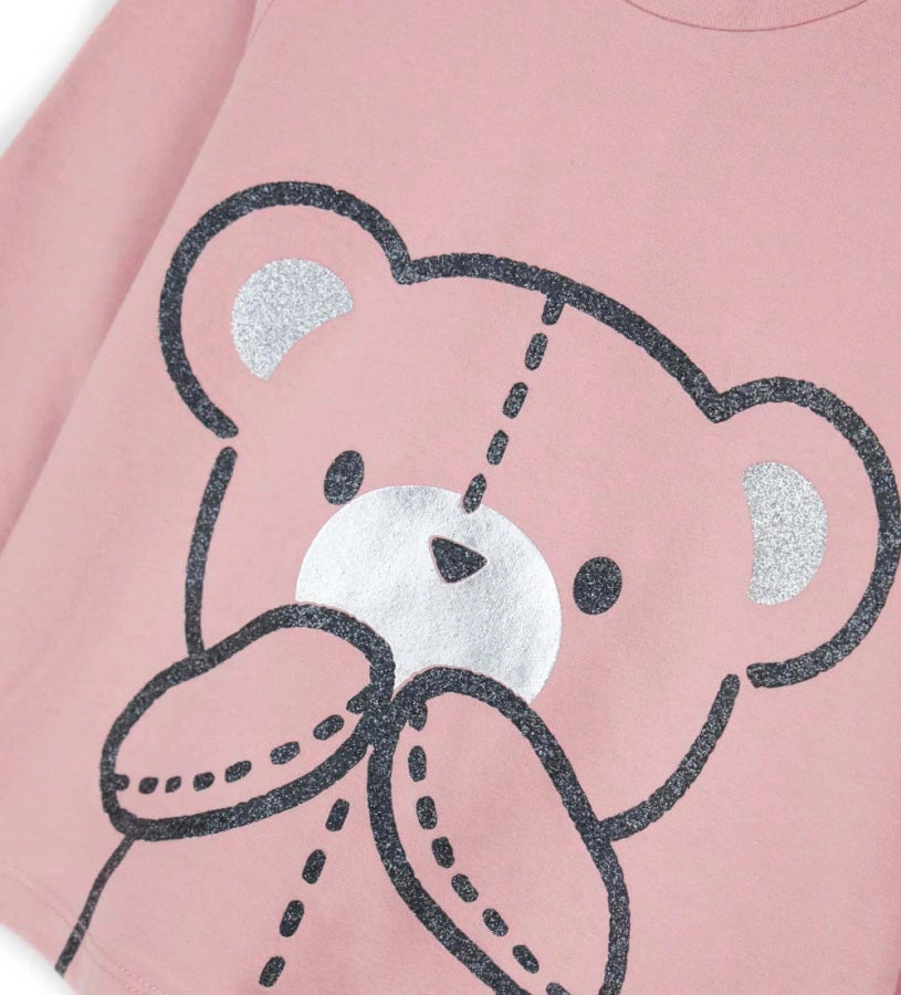 Μπλούζα από ελαστικό βαμβάκι με στρογγυλή λαιμόκοψη, μακριά μανίκια, βολάν στους ώμους και τύπωμα με γκλίτερ και μεταλλικές λεπτομέρειες στο μπροστινό μέρος. Για τα μεγέθη έως 2–3 ετών διαθέτει πρακτικό άνοιγμα με κουμπιά.