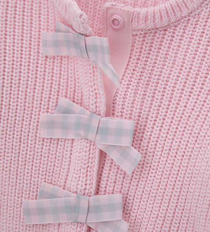 Παιδική ζακέτα από βαμβακερό νήμα με στρογγυλή λαιμόκοψη, ριμπ τελειώματα και μακριά μανίκια. Διαθέτει μπροστινό άνοιγμα με κρυφά κουμπιά-σούστες, διακοσμητικούς φιόγκους από καρό ύφασμα και μενταγιόν στο μπροστινό μέρος. Ένα εκλεπτυσμένο και απαλό κομμάτι, ιδανικό για κοριτσίστικες εμφανίσεις με ρομαντικό στυλ.