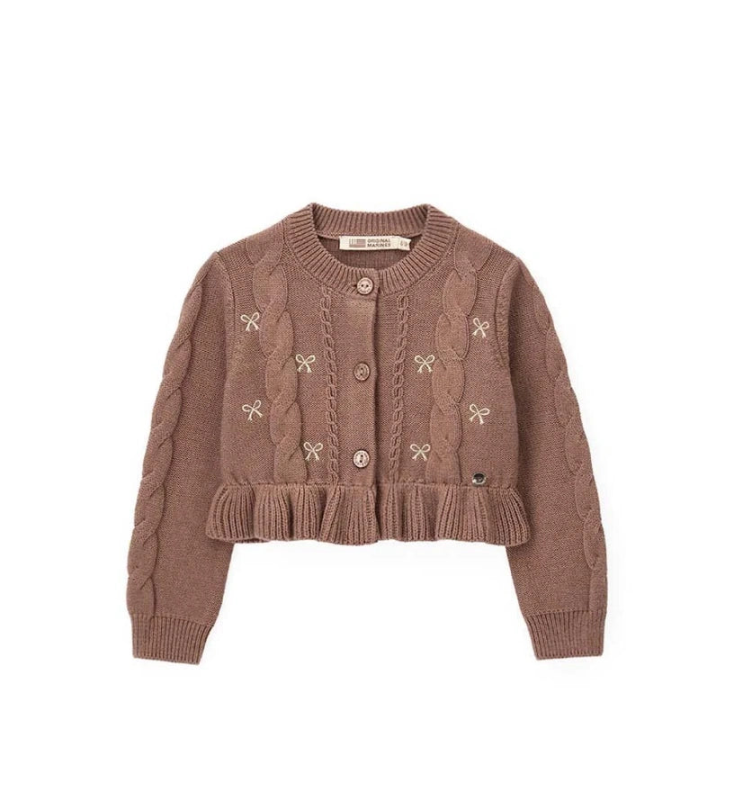 Παιδική ζακέτα από βαμβακερό νήμα με στρογγυλή λαιμόκοψη, ριμπ τελειώματα και μακριά μανίκια. Διαθέτει μπροστινό άνοιγμα με κουμπιά, πλέξη σε σχέδιο πλεξούδας, διακοσμητικό βολάν στο κάτω μέρος και μενταγιόν στο μπροστινό μέρος. Ιδανική για κομψές και απαλές κοριτσίστικες εμφανίσεις με ρομαντικό ύφος.