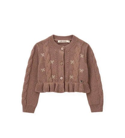 Παιδική ζακέτα από βαμβακερό νήμα με στρογγυλή λαιμόκοψη, ριμπ τελειώματα και μακριά μανίκια. Διαθέτει μπροστινό άνοιγμα με κουμπιά, πλέξη σε σχέδιο πλεξούδας, διακοσμητικό βολάν στο κάτω μέρος και μενταγιόν στο μπροστινό μέρος. Ιδανική για κομψές και απαλές κοριτσίστικες εμφανίσεις με ρομαντικό ύφος.