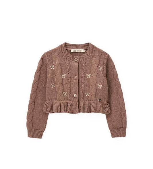 Παιδική ζακέτα από βαμβακερό νήμα με στρογγυλή λαιμόκοψη, ριμπ τελειώματα και μακριά μανίκια. Διαθέτει μπροστινό άνοιγμα με κουμπιά, πλέξη σε σχέδιο πλεξούδας, διακοσμητικό βολάν στο κάτω μέρος και μενταγιόν στο μπροστινό μέρος. Ιδανική για κομψές και απαλές κοριτσίστικες εμφανίσεις με ρομαντικό ύφος.