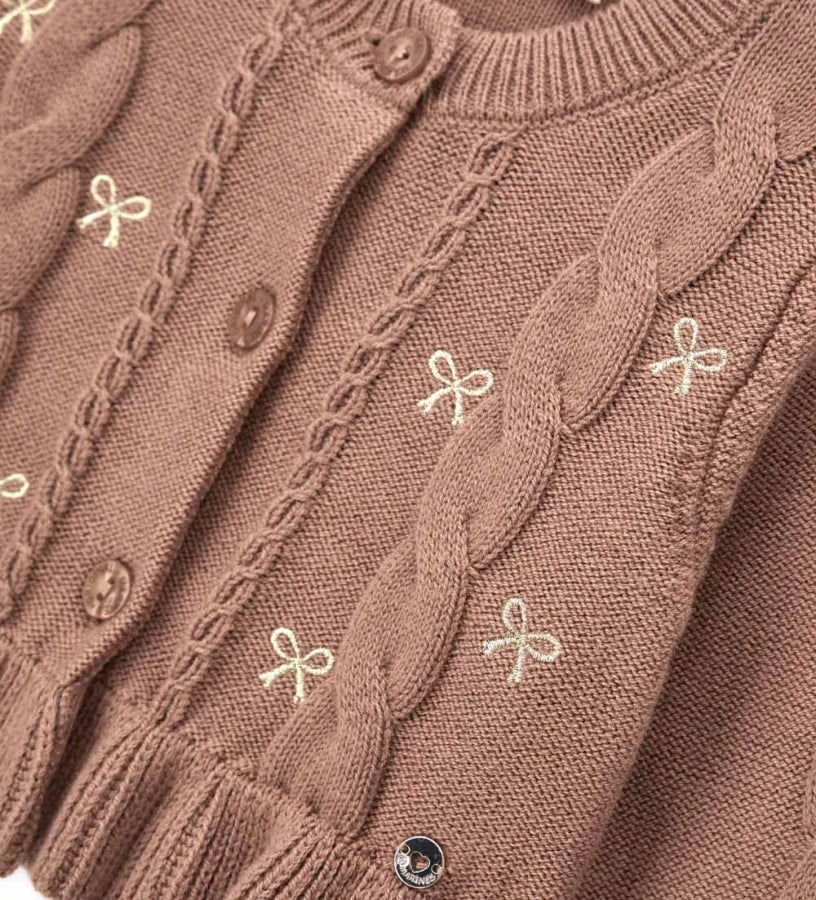 Παιδική ζακέτα από βαμβακερό νήμα με στρογγυλή λαιμόκοψη, ριμπ τελειώματα και μακριά μανίκια. Διαθέτει μπροστινό άνοιγμα με κουμπιά, πλέξη σε σχέδιο πλεξούδας, διακοσμητικό βολάν στο κάτω μέρος και μενταγιόν στο μπροστινό μέρος. Ιδανική για κομψές και απαλές κοριτσίστικες εμφανίσεις με ρομαντικό ύφος.