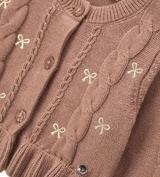 Παιδική ζακέτα από βαμβακερό νήμα με στρογγυλή λαιμόκοψη, ριμπ τελειώματα και μακριά μανίκια. Διαθέτει μπροστινό άνοιγμα με κουμπιά, πλέξη σε σχέδιο πλεξούδας, διακοσμητικό βολάν στο κάτω μέρος και μενταγιόν στο μπροστινό μέρος. Ιδανική για κομψές και απαλές κοριτσίστικες εμφανίσεις με ρομαντικό ύφος.