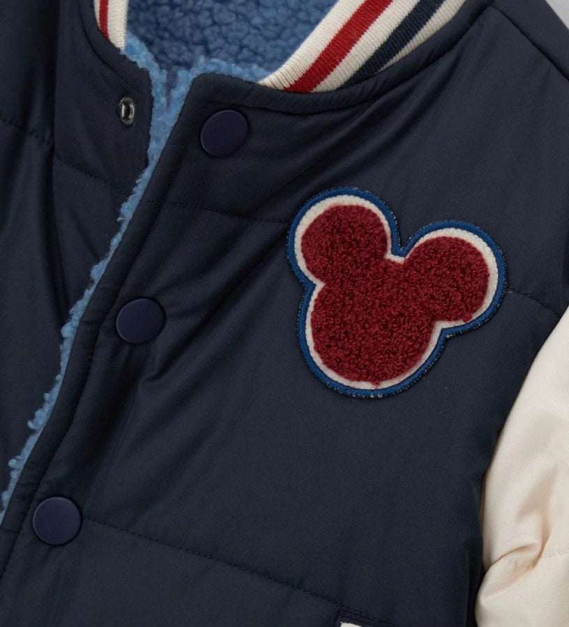 Αναστρέψιμο μπουφάν bomber Disney Mickey Mouse με γέμισμα. Η μία πλευρά από νάιλον με μανίκια σε αντίθεση και μπροστινό απλικέ, και η δεύτερη πλευρά από ζεστό οικολογικό teddy με μανίκια σε αντίθεση και απλικέ. Κλείσιμο με κουμπιά-σούστες και ριμπ τελειώματα από βαμμένο νήμα για ένα δυναμικό και ζεστό στυλ.